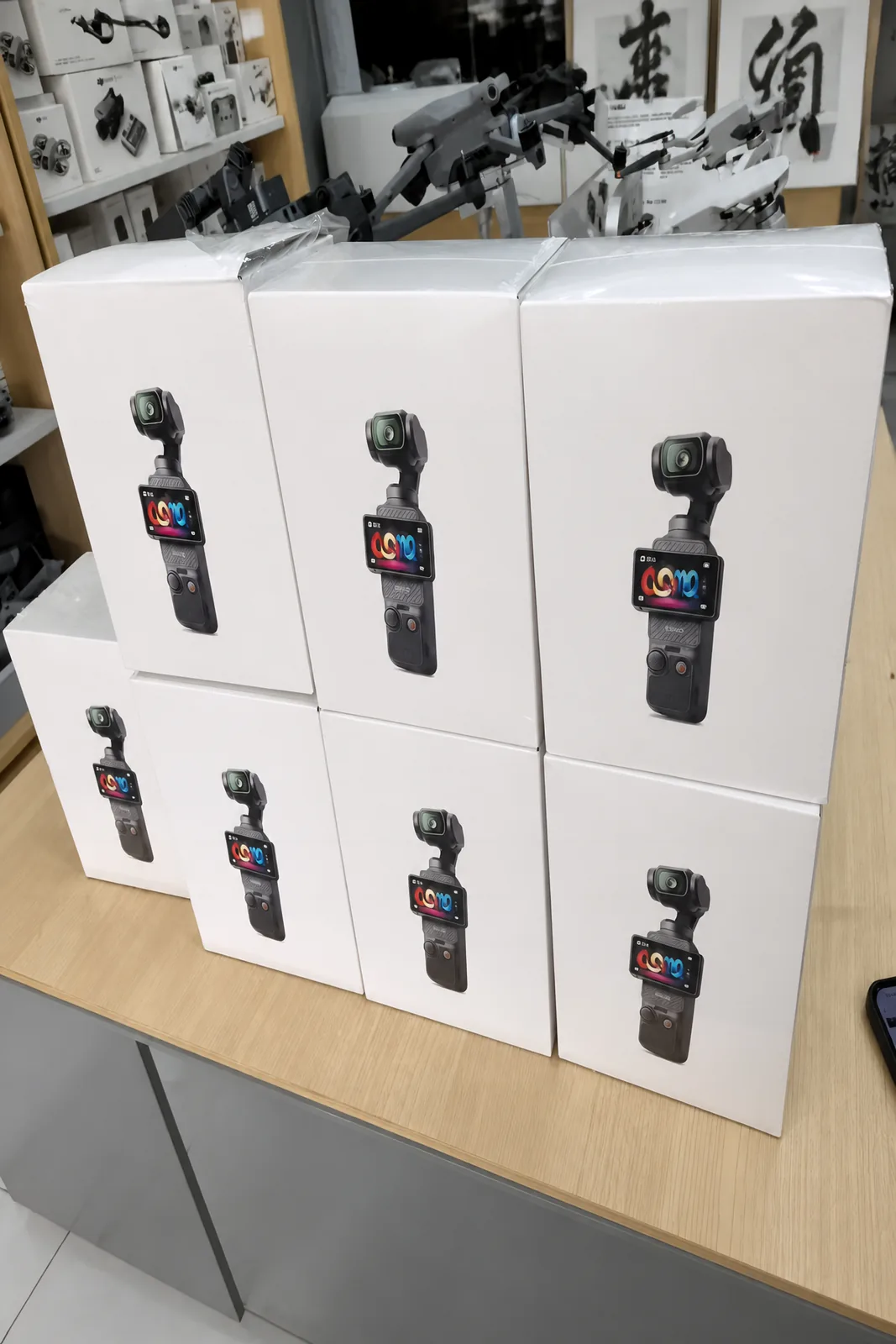 Boxed DJI Osmo Pocket 3 cameras stacked on a desk, with drones on the shelves behind